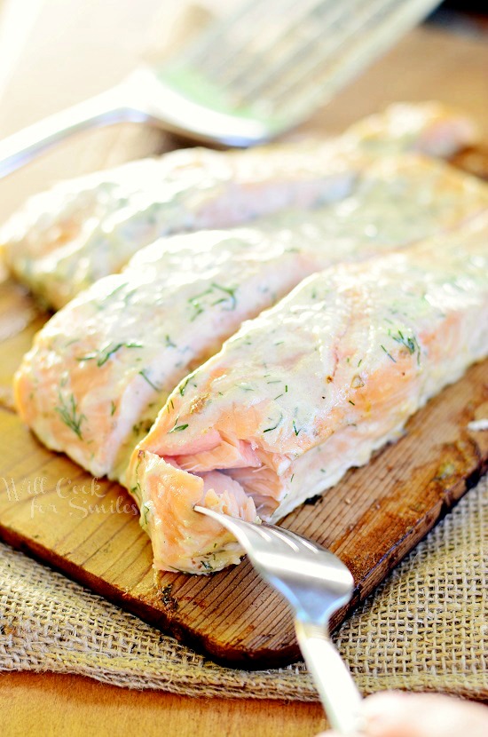 cooked Cedar Plank Salmon smothered in creamy lemon dill sauce on a cutting board with a bite on a fork and a spatula in the background 
