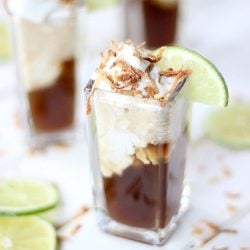clear square shaped glass filled with dirty dr. pepper floats on a white table with lime slices to left and additional floats in background