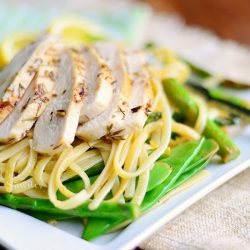 lemon herb pasta on a square white plate with lemon garnish on a brown cloth on top of wood table