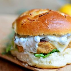 Salmon burger on a bun on wood plank with white sauce on patty with a lemon in the background