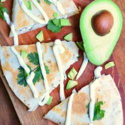 above view of avocado chicken quesadilla on a wood cutting board on wood table with slice avocado in background while quesadilla is drizzled with sour cream