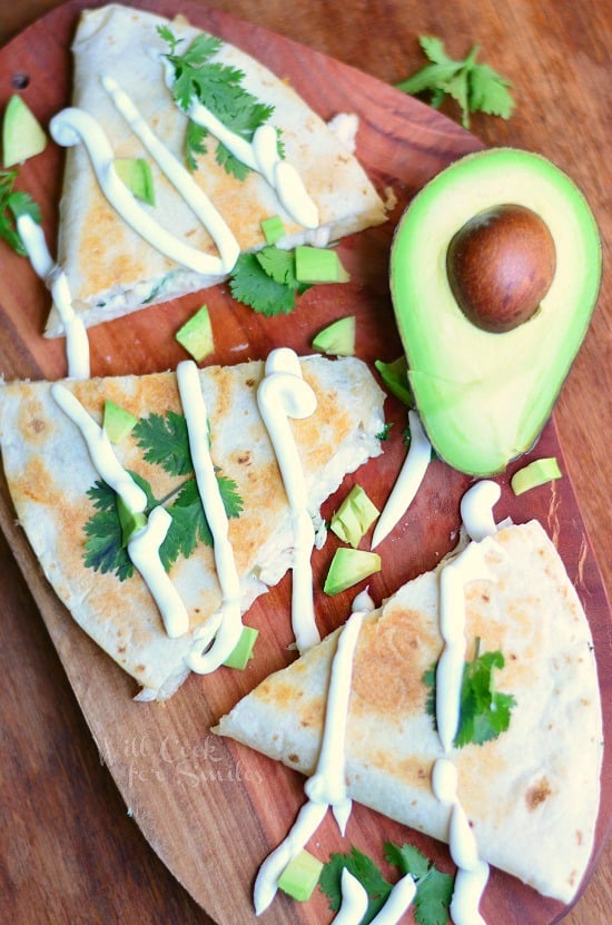 above view of avocado chicken quesadilla on a wood cutting board on wood table with slice avocado in background while quesadilla is drizzled with sour cream