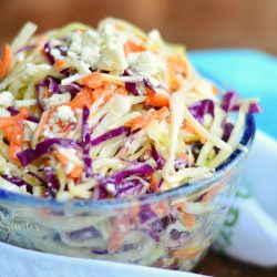 close up view of clear glass bowl filled with blue cheese coleslaw on a wood table with a white and green cloth in foreground and a blue cloth in background