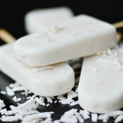 close up view of 3 coconut vanilla creamy pops on a black table with shredded coconut strewn across pops and table