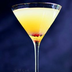 front view of martini glass filled with flirtini pineapple champagne martini on a black table with blue background