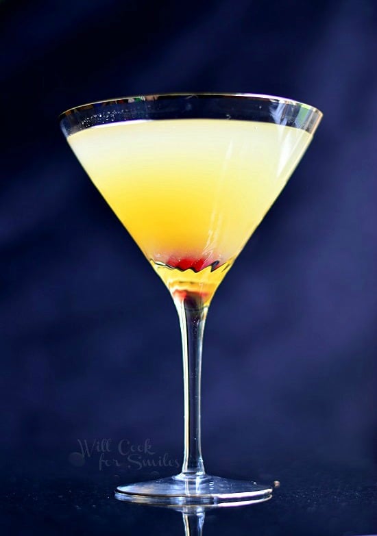 Flirtini is in a tall martini glass. The liquid is musky yellow with red in the bottom of the glass from the cherries.