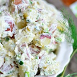 view from above of close up view of white decorative bowl filled with heaping portion of Parmesan Herb Potato salad on a wood board with cloth in background