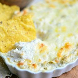 chip in dip in small white baking dish filled with chicken enchilada verde dip on a wood table with tan cloth to the left and a bowl of chips in background