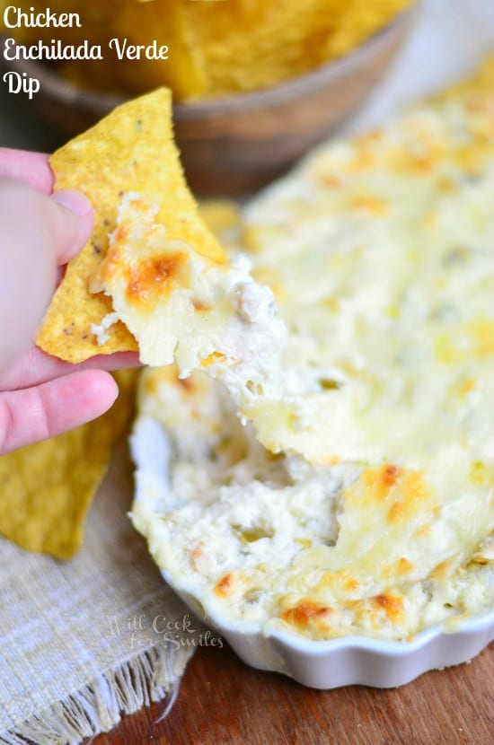 Chicken Enchilada Verde Dip is presented in a white dish. The left hand holds a tortilla chip that has been dipped into the Chicken Enchilada Verde Dip. A wooden bowl containing tortilla chips fades into the background,