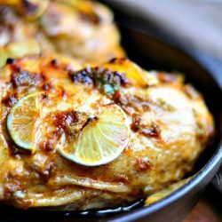 view from close up Black skillet with chili lime roasted chicken with sliced lime on top while skillet is on a wooden table with white and grey cloths around