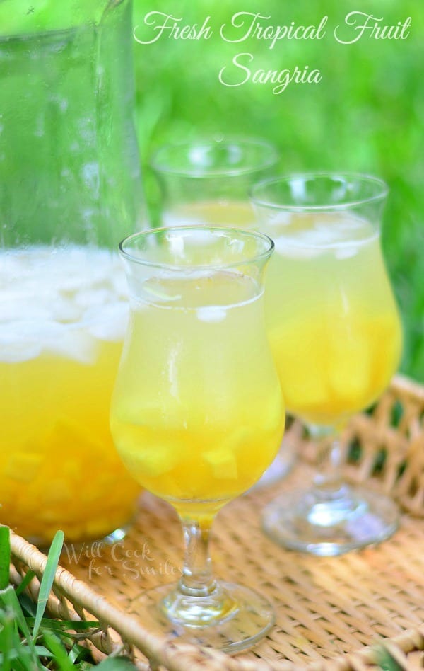 3 wine glasses filled with fresh tropical fruit sangria with a glass pitcher half filled with the sangria on a wicker serving tray with sweetnlow packets below glasses.