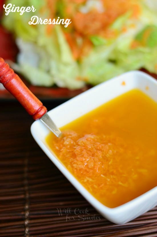 Ginger Dressing is presented in a white, square bowl. It's dark yellow in color. There are small pieces of grated carrot and ginger seen within the dressing.