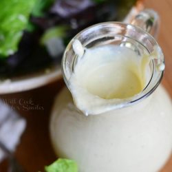 close up from above of small dressing jar filled with light caesar dressing on a wood table in fron of a ceramic bowl filled with caesar salad