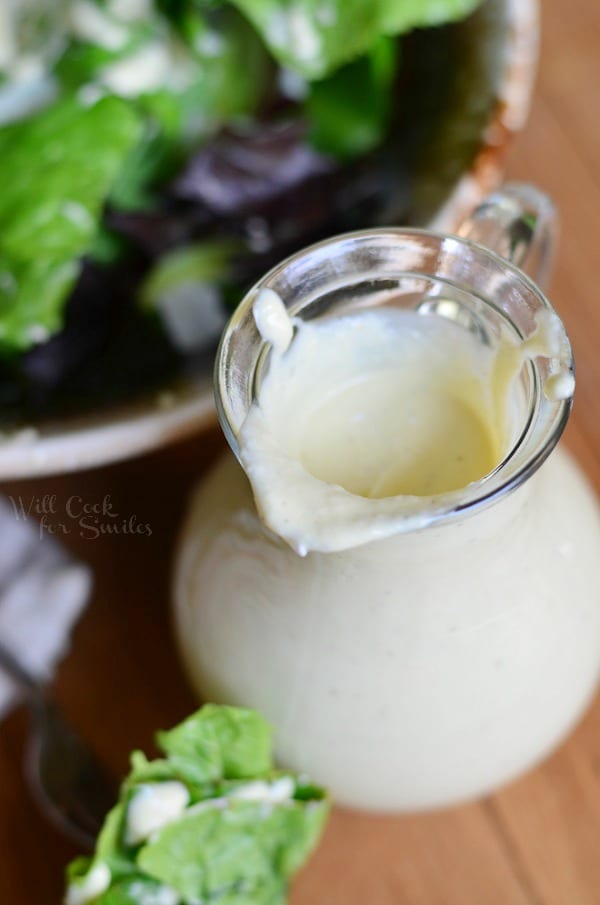 close up from above of small dressing jar filled with light caesar dressing on a wood table in fron of a ceramic bowl filled with caesar salad