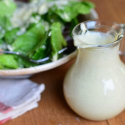 small dressing jar filled with light caesar dressing on a wood table in fron of a ceramic bowl filled with caesar salad