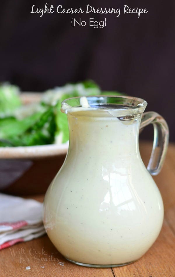 Light Caesar Dressing Recipe {No Egg} is served in a pouring dish. It's white in color. There is a bowl of salad behind the dressing that fades into the background.