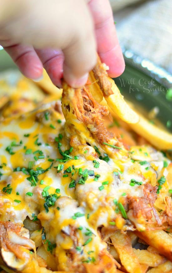 hand pulling out fries from pulled pork loaded fries on a silver platter covered in scallions and mixed melted cheese
