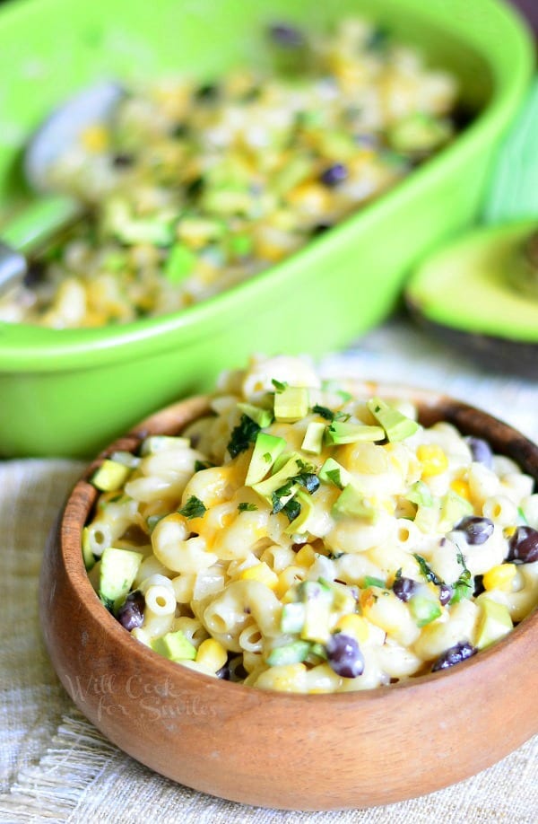 Tex-Mex Mac and Cheese in a wood bowl with avocado on top and the rest of mac and cheese in a green casserole dish 