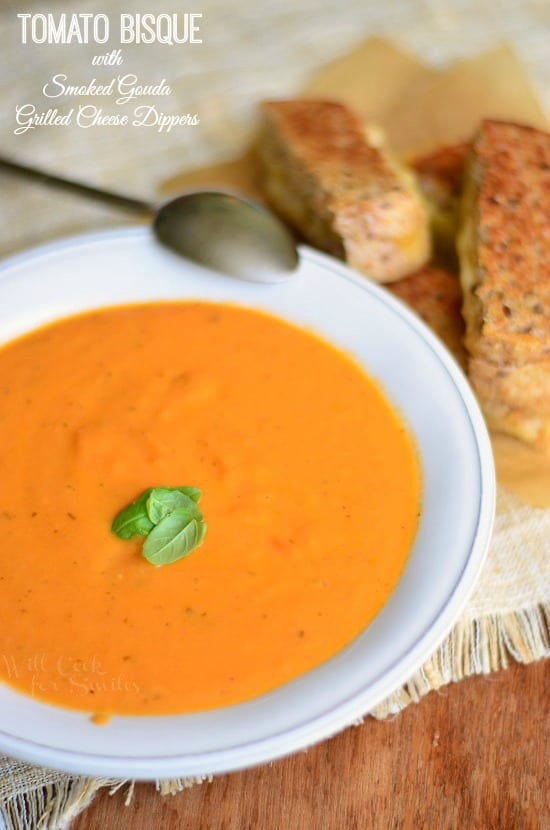 view from above of tomato bisque in a white bowl with grey rim with a spoon leaning on top of bowl and smoked gouda grilled cheese dippers to the top right of bowl