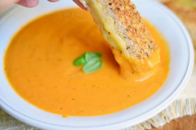 Tomato Bisque in a white bowl with basil on top and with Grilled Cheese Dipper being dipped in it 