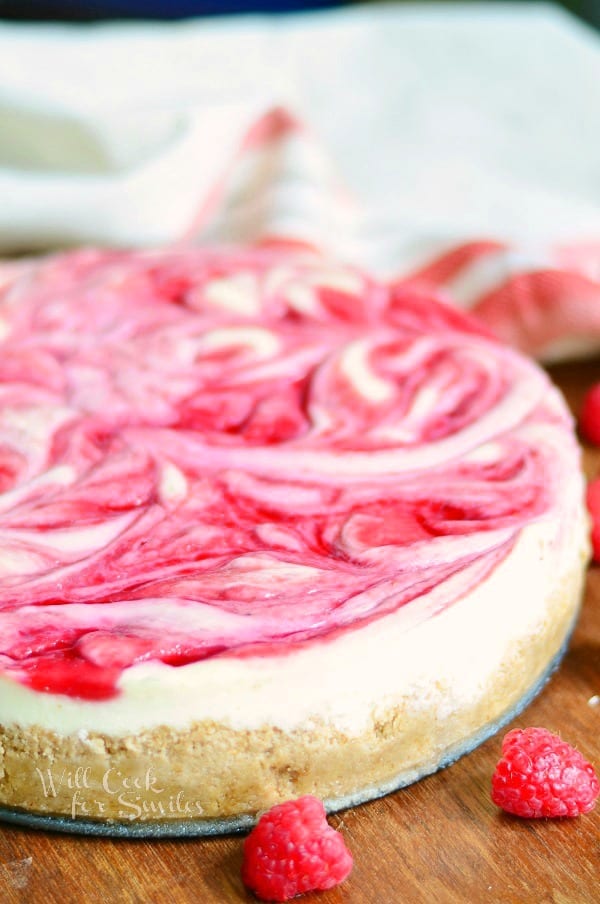 whole white chocolate raspberry swirl cheesecake on a wooden table with 2 raspberries in foreground and white cloth behind cake in background