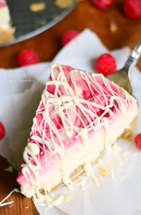 above view of 1 slice of white chocolate raspberry swirl cheesecake with berries around slice and whole cake in oan above to the left