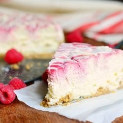 1 slice of white chocolate raspberry swirl cheesecake on a wooden table with 2 raspberries in foreground and whole cake and white cloth behind cake in background
