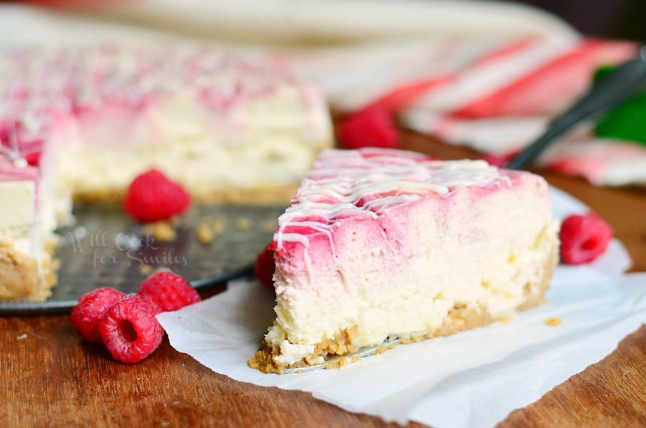 1 slice of white chocolate raspberry swirl cheesecake on a wooden table with 2 raspberries in foreground and whole cake and white cloth behind cake in background