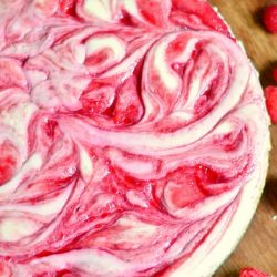 whole white chocolate raspberry swirl cheesecake on a wooden table with 2 raspberries in foreground and white cloth behind cake in background