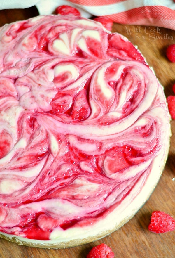 whole white chocolate raspberry swirl cheesecake on a wooden table with 2 raspberries in foreground and white cloth behind cake in background