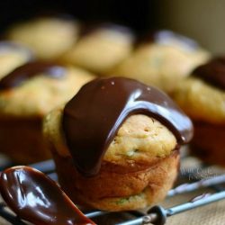 zucchini muffins on a cooling rack sitting on a burlap placemat on wooden table with spoon with hot fudge topping leaning on rack