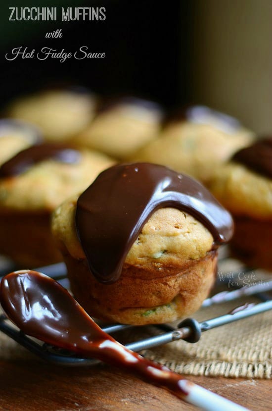 zucchini muffins on a cooling rack sitting on a burlap placemat on wooden table with spoon with hot fudge topping leaning on rack