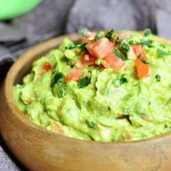 close up view wooden bowl filled with Guacamole topped with tomatoes and cilantro on a wooden table with blue tortilla chips in background around bowl