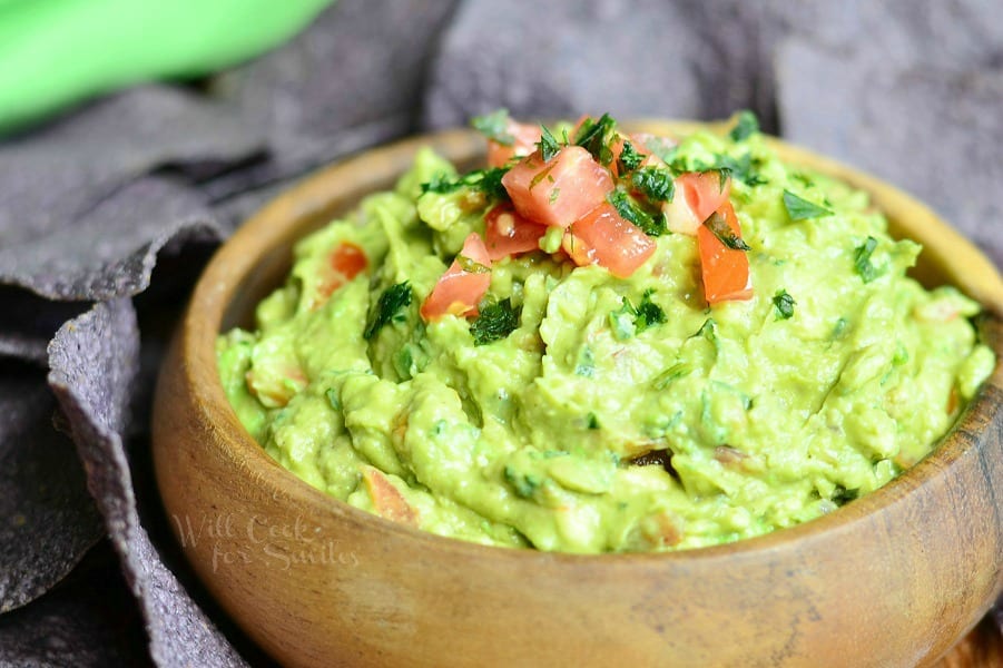 Guacamole in a wood bowl with tomato on top for garnish and chips around it 