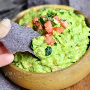 hand scooping guacamole with chip out of wooden bowl filled with Guacamole topped with tomatoes and cilantro on a wooden table with blue tortilla chips in background around bowl