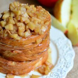 close up of 3 portions of apple pie stuffed french toast topped with baked apples on white plate sitting on top of brown placemat with sliced apples in background