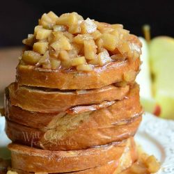 3 portions of apple pie stuffed french toast topped with baked apples on white plate sitting on top of brown placemat with sliced apples in background