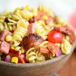 close up view of meat lovers pasta salad in a wooden bowl on a wood table with white and red cloth in background