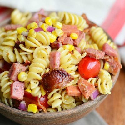 above view and close up view of meat lovers pasta salad in a wooden bowl on a wood table with white and red cloth in background