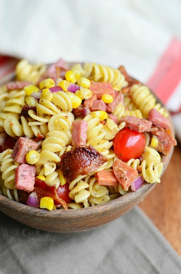 above view and close up view of meat lovers pasta salad in a wooden bowl on a wood table with white and red cloth in background