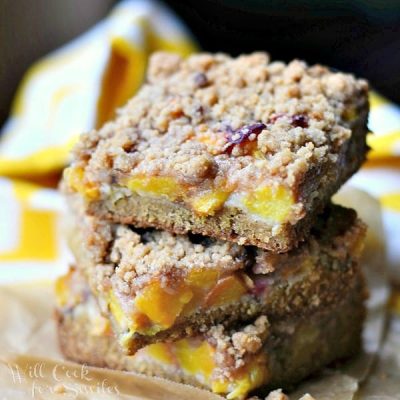 3 peach crumble cookie bars stacked on a brown piece of wax paper on a wooden table with a yellow and white cloth in background