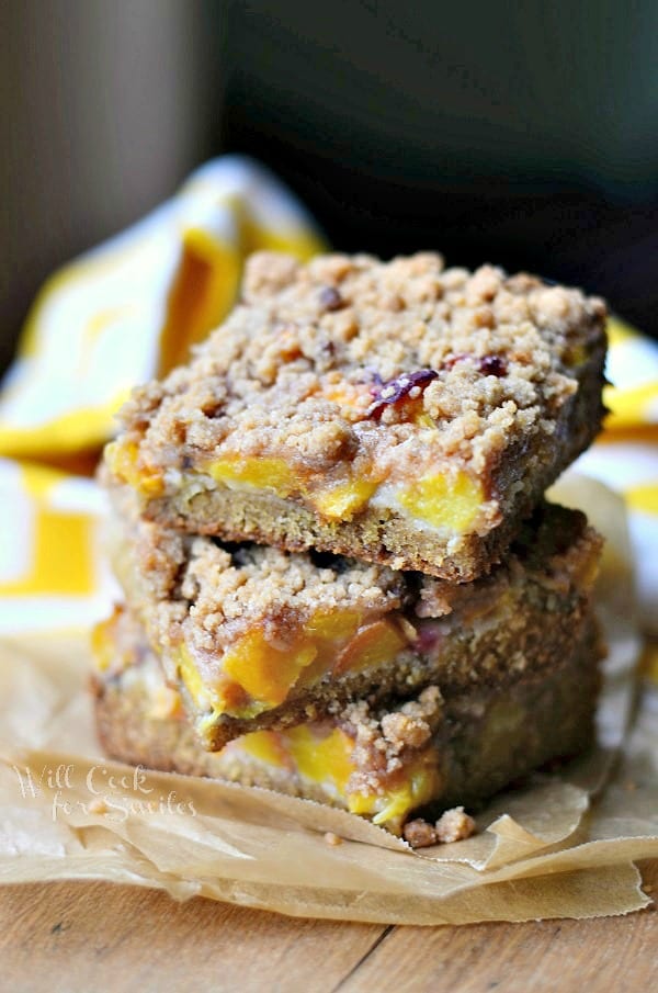 3 peach crumble cookie bars stacked on a brown piece of wax paper on a wooden table with a yellow and white cloth in background