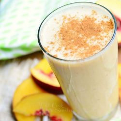 glass pint glass filled with peaches and cream breakfast smootie on a brown placemat on wood table with slices of peach at base of glass