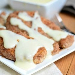 3 pecan crusted chicken tenders covered in cheese sauce on a white rectangular plate on wood table with white cloth and decorative fork below plate. Square bowl in background with additional sauce.