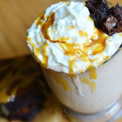 view from above of Glass mug filled with salted caramel brownie milkshake on a wooden table with a brownie in front of glass while both are drizzled with caramel