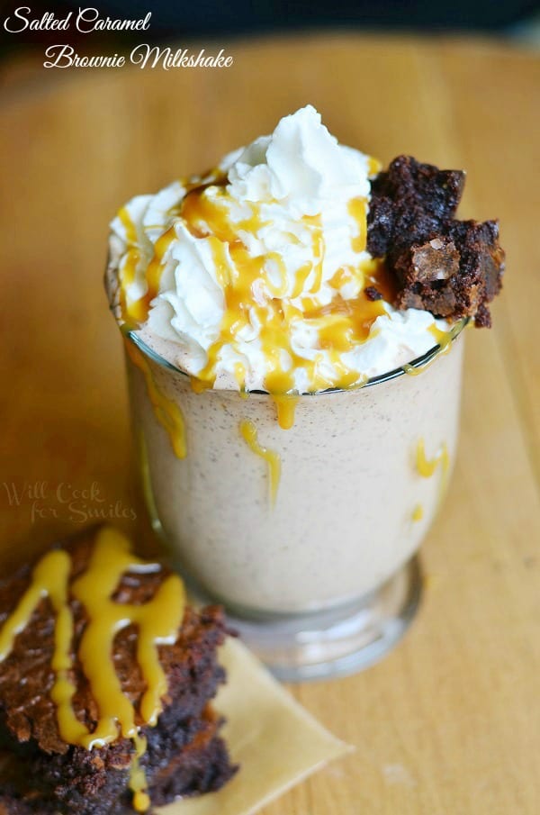 Glass mug filled with salted caramel brownie milkshake on a wooden table with a brownie in front of glass while both are drizzled with caramel