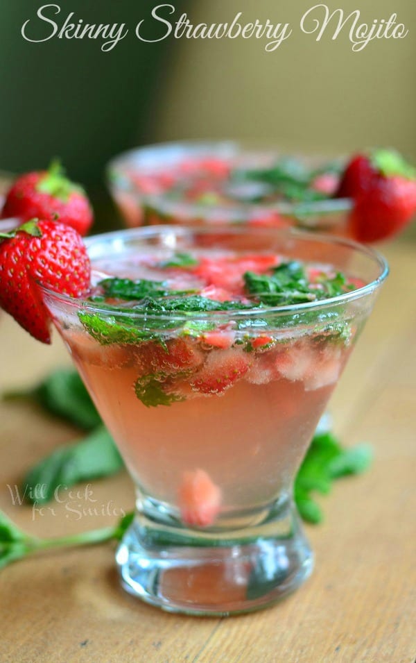 Skinny Strawberry Mojito is presented in a short martini glass. There are pieces of strawberry and mint floating in the glass. A strawberry sits on each rim as well. Mint lays on the table.
