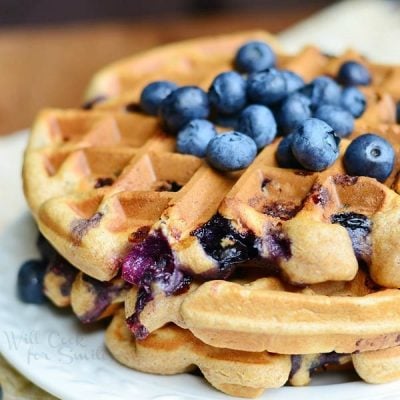 round white plate with blueberry waffles and blueberries topping the waffles all on a wood table with tan cloth and blueberries below plate