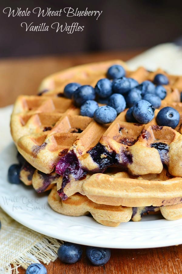 Two Whole Wheat Blueberry Vanilla Waffles stacked on a white plate. Fresh, whole blueberries are on top of the waffles.