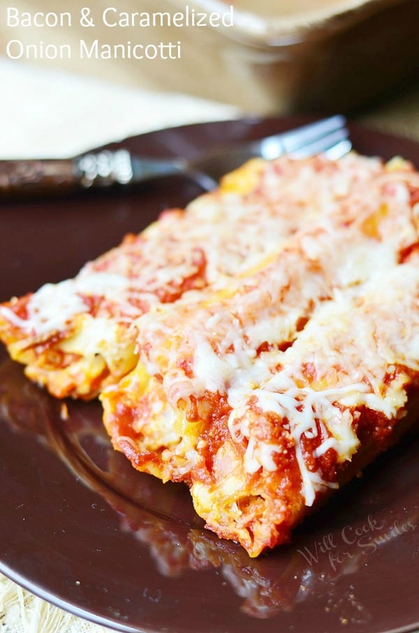3 piecesof bacon caramelized onion manicotti on a black plate with a decorative fork above manicotti on plate on white cloth